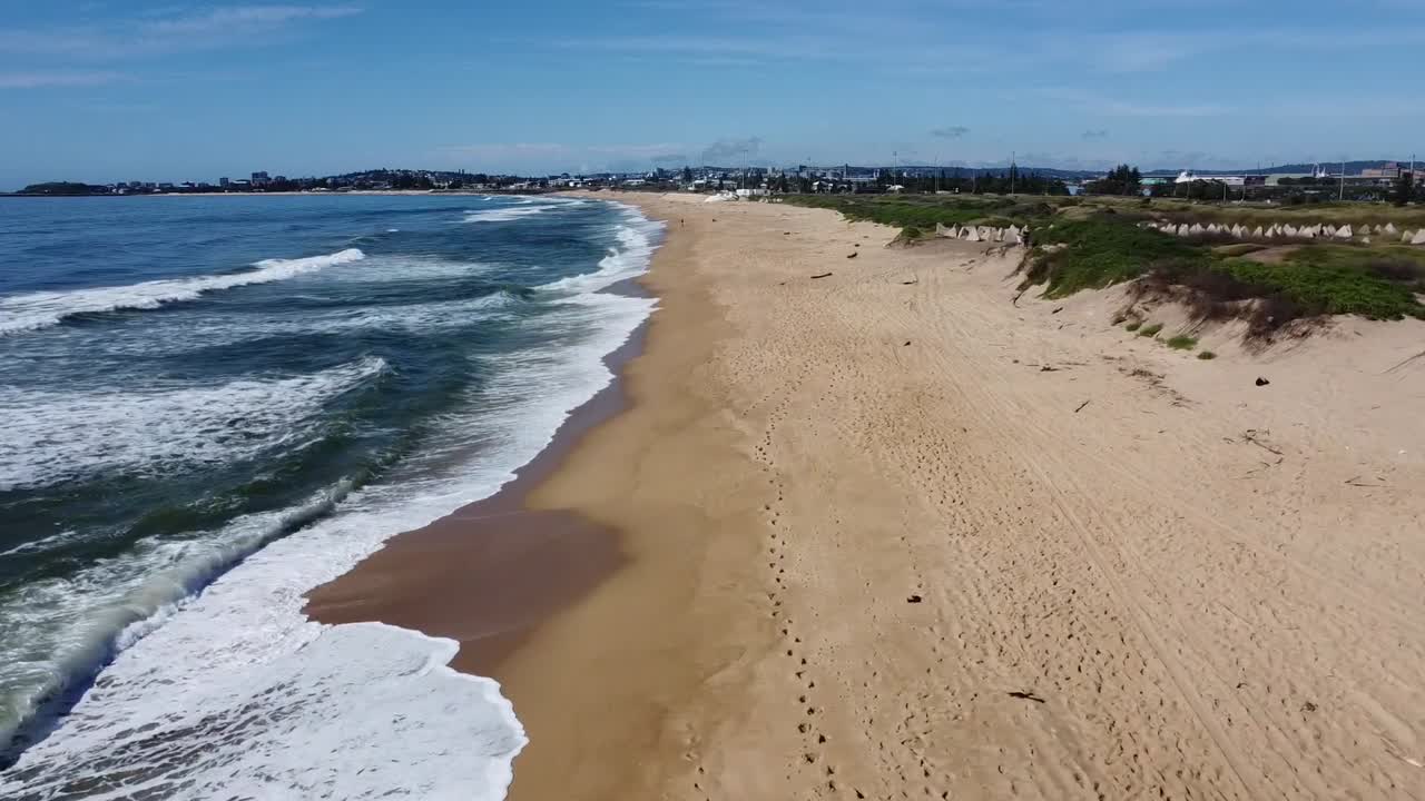 Aerial view of a golden sand beach with coastal dunes and waves gently rolling in, while distant buildings line the horizon, offering a serene and tranquil coastal landscape near Ke Ga