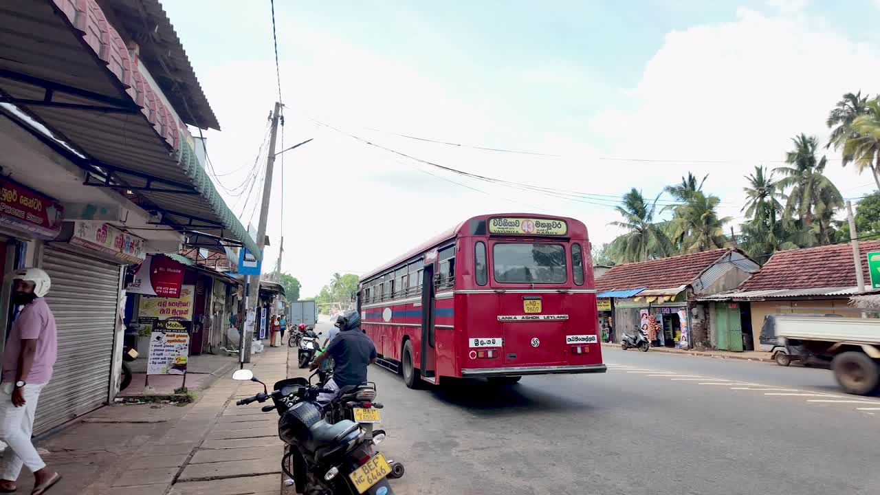 Motorcyclist rides past a parked red bus in a bustling Sri Lankan town, capturing the essence of local transport and daily life.