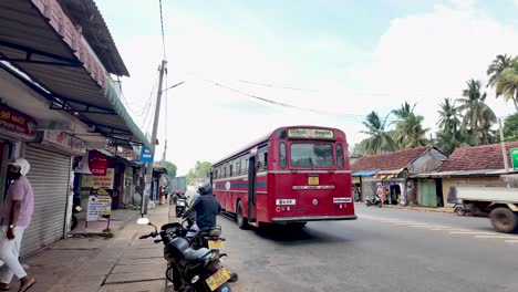 motorcyclist rides past a parked red bus in a bustling sri lankan town, capturing the essence of local transport and daily life.