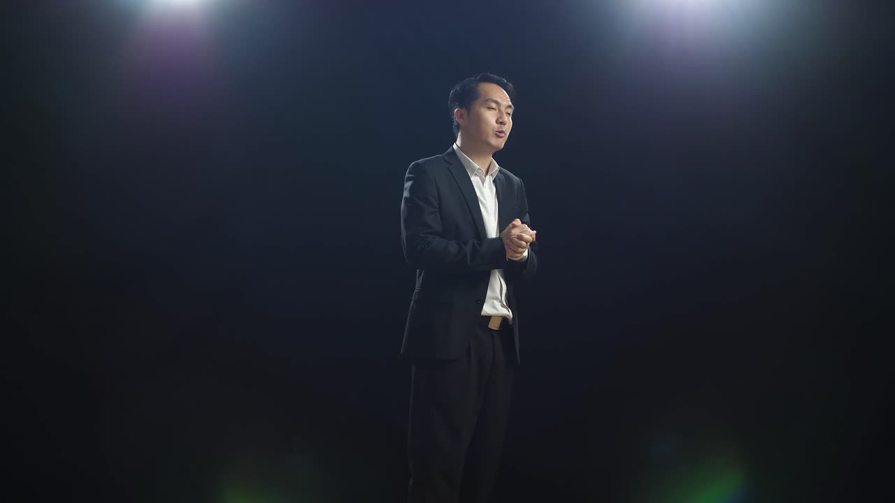 Side View Of Asian Speaker Man In Business Suit Holding His Hands Together And Showing Index Fingers Up While Speaking In The Black Screen Studio