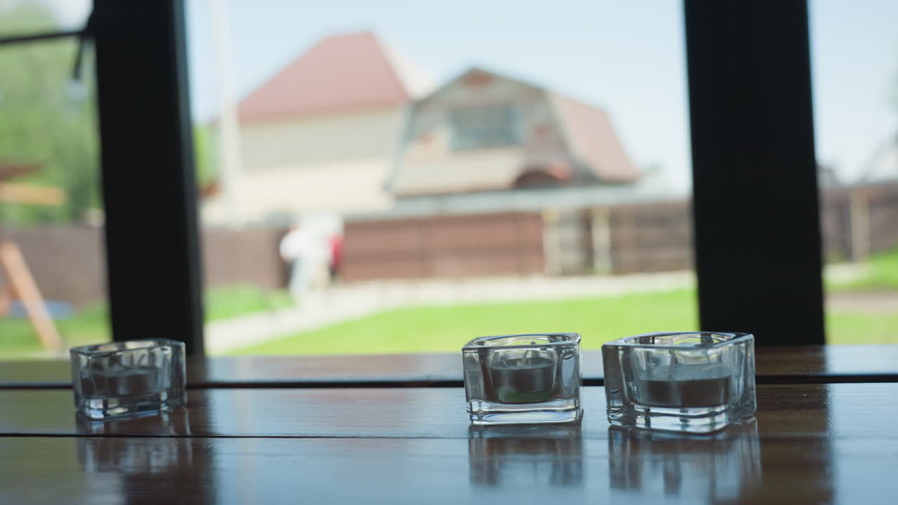 Three small glass candle holders placed on wooden window surface with soft natural light illuminating scene, while blurred figures of people walking in distance create calm background
