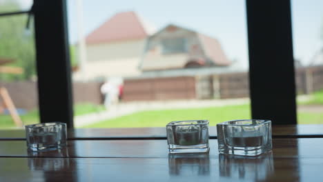 three small glass candle holders placed on wooden window surface with soft natural light illuminating scene, while blurred figures of people walking in distance create calm background