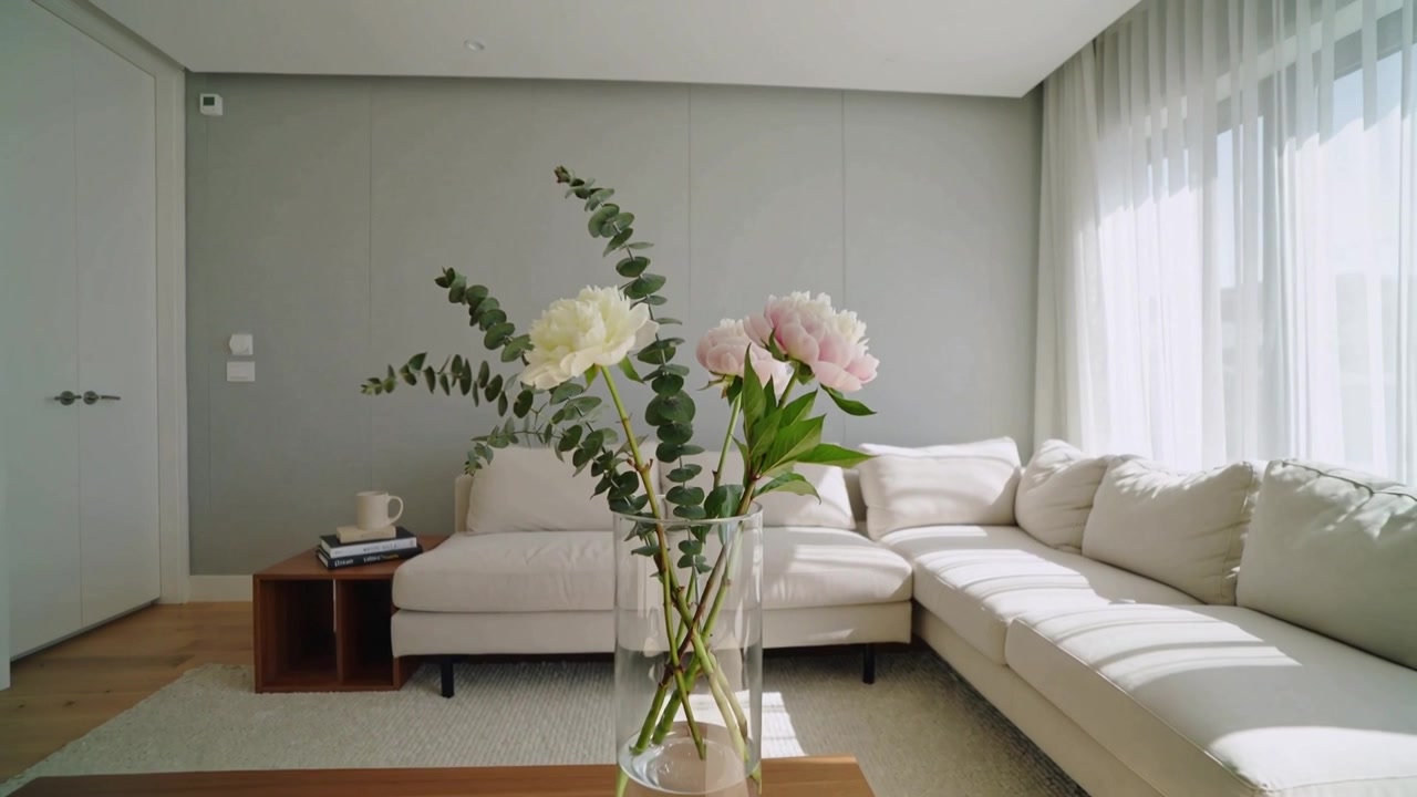 La telecamera si allontana, rivelando un vaso di vetro con eucalipto e peonie su un tavolo in un soggiorno.