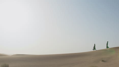 extreme long shot of two muslim women wearing traditional black dress and hijab walking in a windy desert