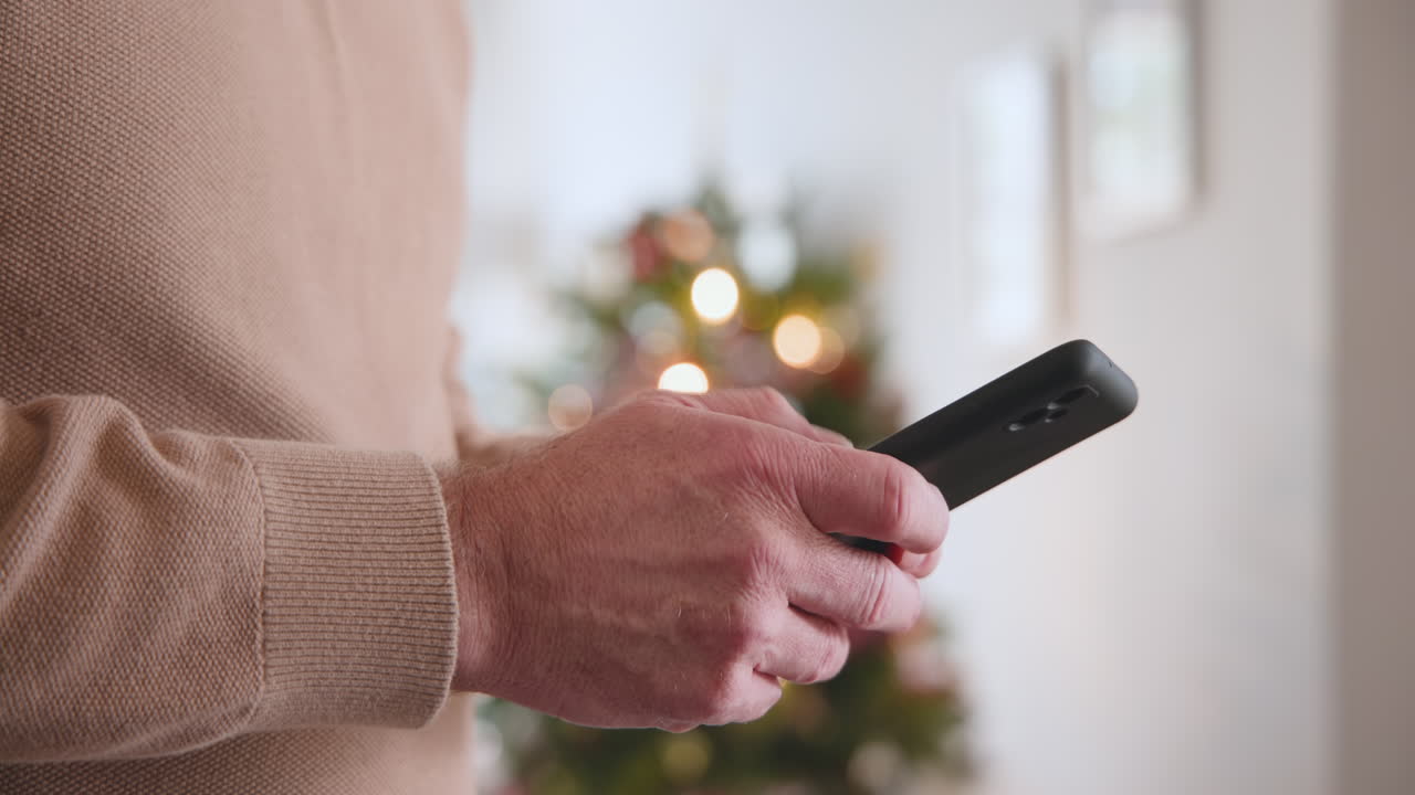 Senior man texting on smartphone near Christmas tree at home