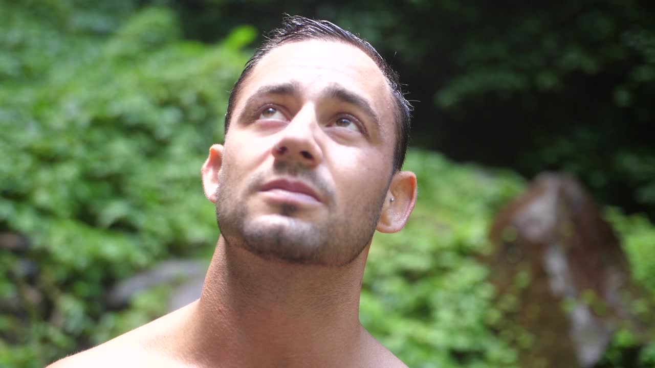 진지한 얼굴 표정을 가진 젊고 매력적인 남자가 카메라를 바라보고 하늘을 향하고 발리의 무성한 녹색 정글에서 주변 환경을 바라보고 있습니다.