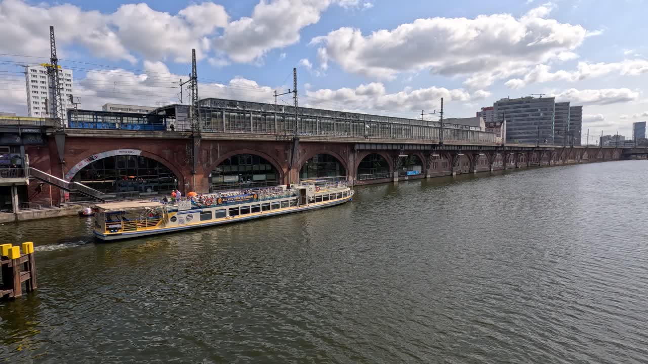 A passenger tour boat glides along a river beneath a historic brick railway bridge in Berlin, with modern city buildings and bright daylight