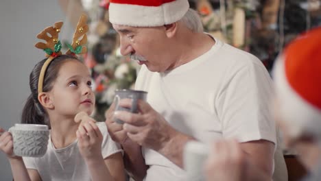 happy grandfather and cute little girl in santa hat and festive headband drinking hot chocolate and eating cookies while chatting with grandmother on christmas eve 1