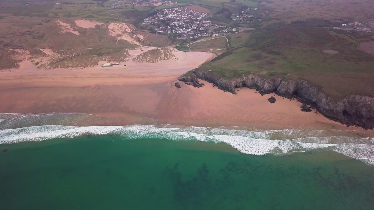 Klidné vlny narážející na širokou písečnou pláž s nedalekou vesnicí v pozadí. Záběr z dronu. Cornwall