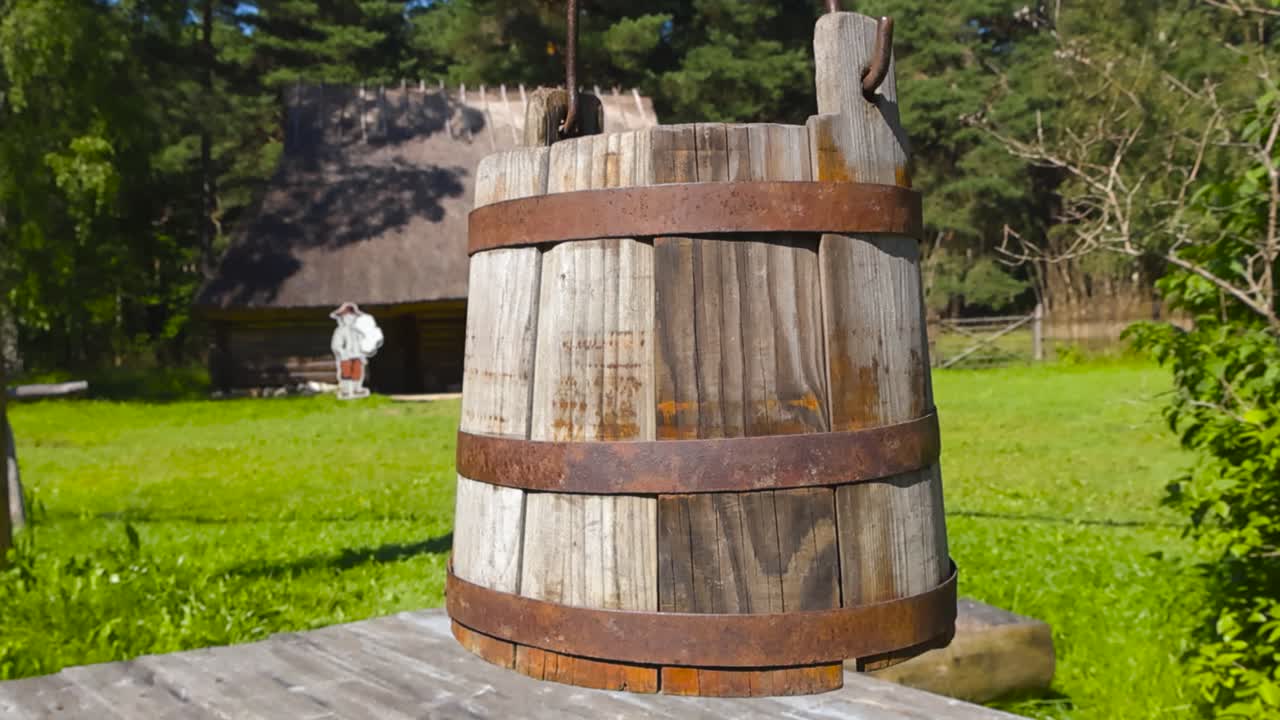 Primer plano con círculos alrededor de un shaduf, un cubo de madera para sacar agua de un pozo que cuelga en el aire. Al fondo, una granja ligeramente desenfocada con césped verde, árboles frondosos y una casa con techo de paja. Jardín rural soleado en verano.