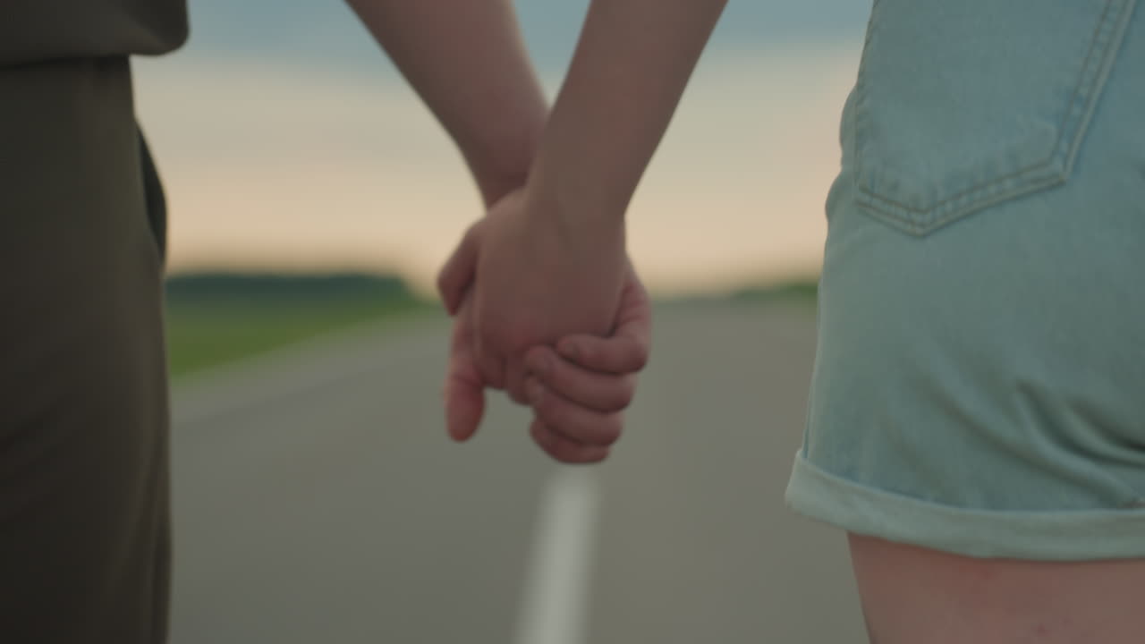 close up rear view of lovers walking hand in hand along empty country road under dramatic sunset clouds with green fields on horizon evoking romance togetherness journey and freedom