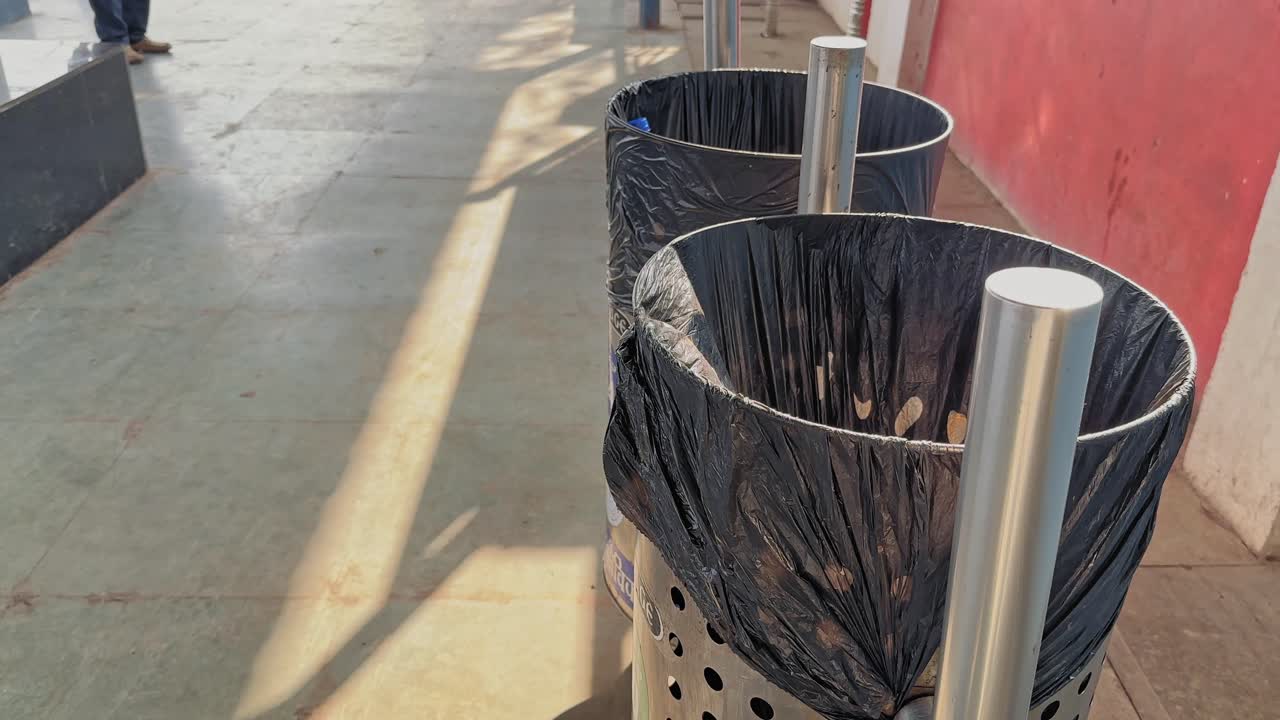 Inquadratura con carrello in avanti su bidoni della spazzatura in acciaio inossidabile posizionati lungo il camminamento della piattaforma ferroviaria, che mostra l'installazione per la raccolta dei rifiuti con luce solare e ombre sul pavimento della stazione