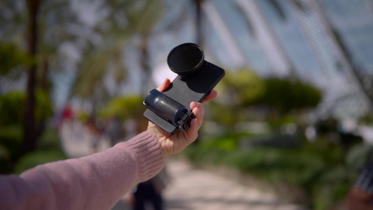 Primer plano de una mano extendiendo un smartphone equipado con una empuñadura estabilizadora compacta y un filtro oscuro circular, presentado hacia adelante en una demostración estable al aire libre en medio de un sendero de parque soleado y vegetación suave