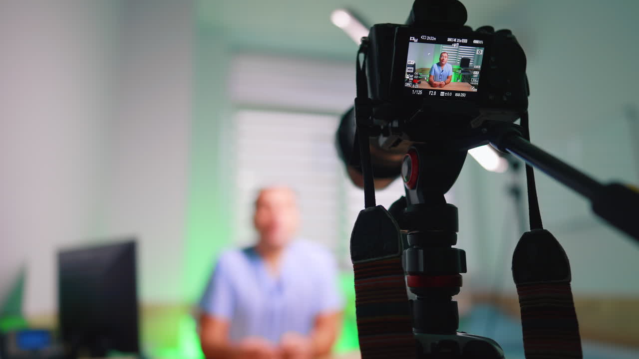 Vista da telecamera posteriore di medico che registra video medico. Vista da telecamera posteriore che riprende un medico in camice blu, con retroilluminazione verde della clinica e tapparelle sfocate.