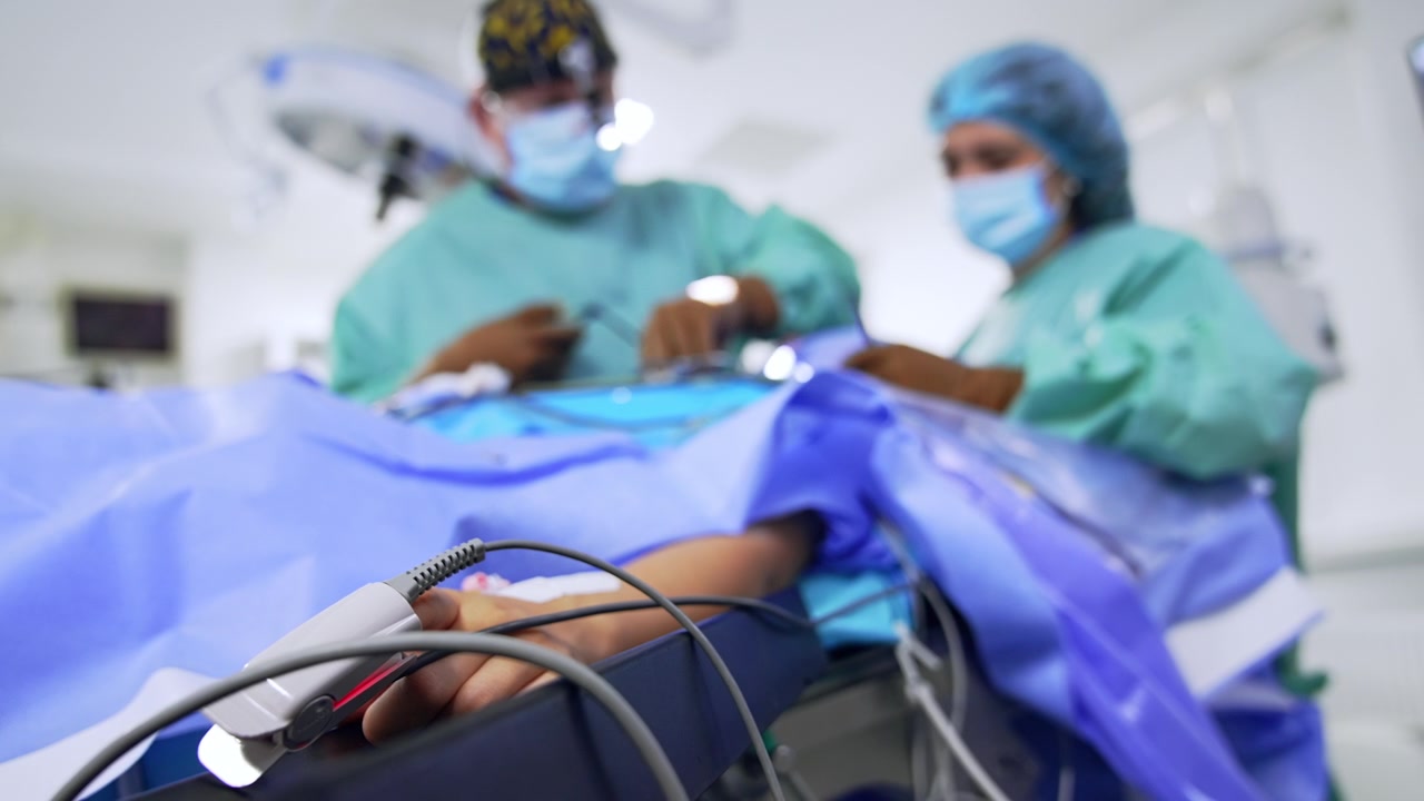 Sensore sulla mano di un paziente sotto intervento. Primo piano. Chirurgo uomo e infermiera donna eseguono un'operazione al naso. Vista dal basso.