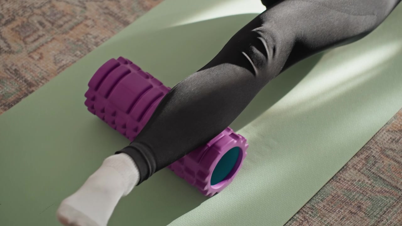 Home mat foam roller under foot during after run recovery, focus on plantar and ankle mobility, purple textured roller on green mat, socks and leggings, slow pressure rolling for relief
