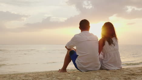 Ein-Mann-Und-Eine-Frau-Sitzen-Nebeneinander-Im-Sand-Am-Strand-Zusammen-Schauen-Sie-Sich-Den-Sonnenuntergang-An