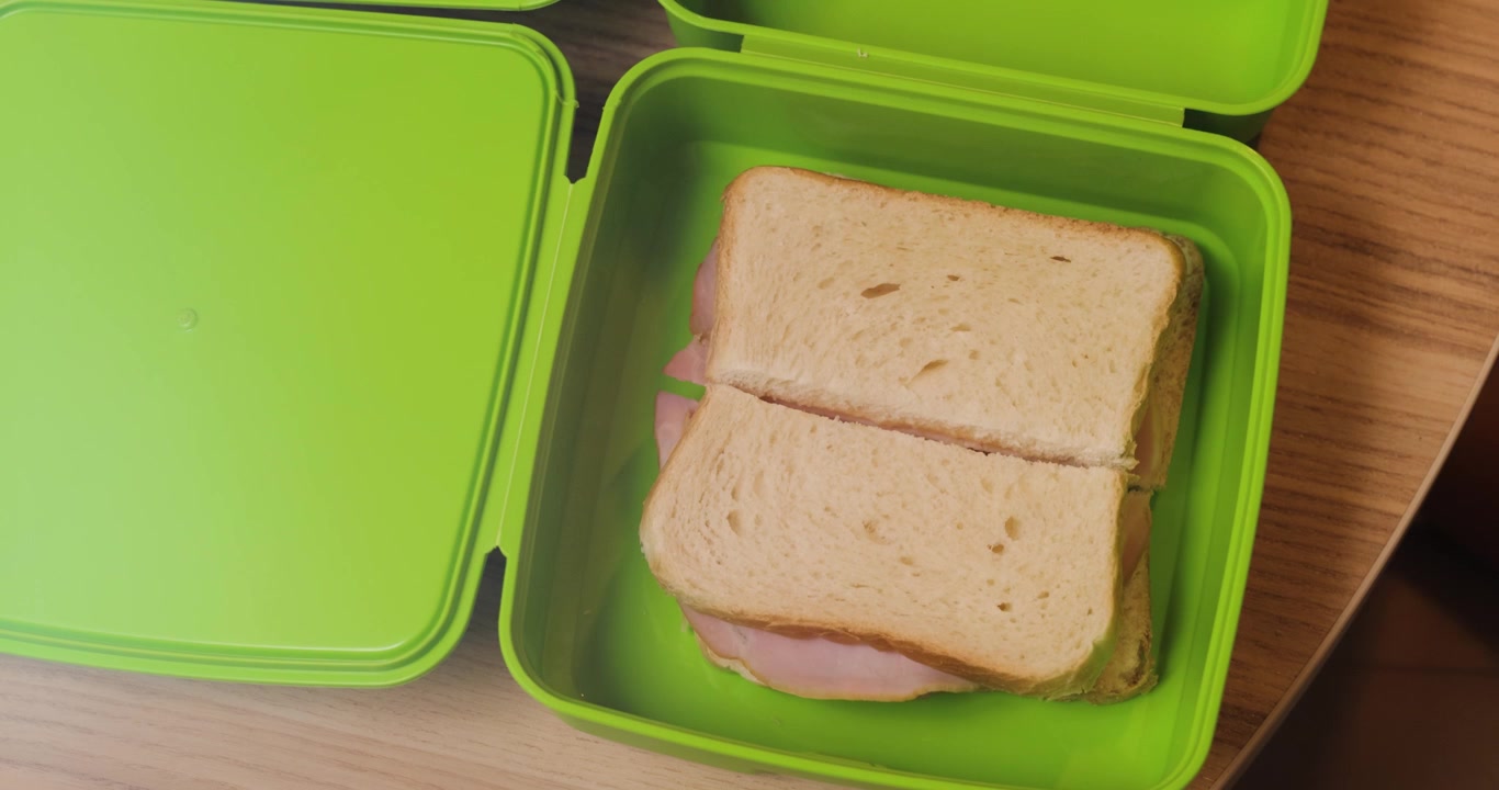 Primer plano de la mano de una mujer joven colocando sándwiches de jamón en una fiambrera verde, en resolución 4K