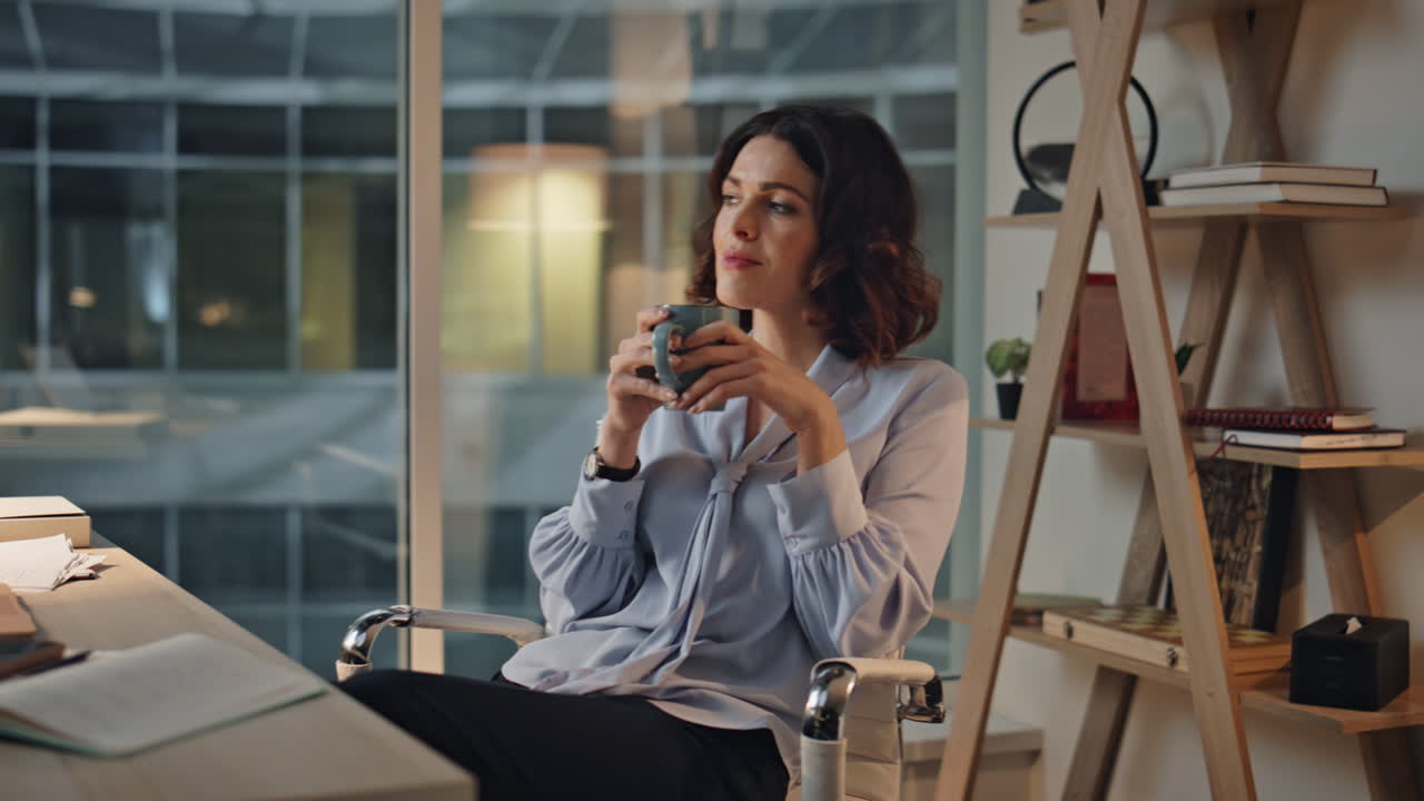 Thoughtful manager sipping tea sitting at office desk at night. Woman drinking