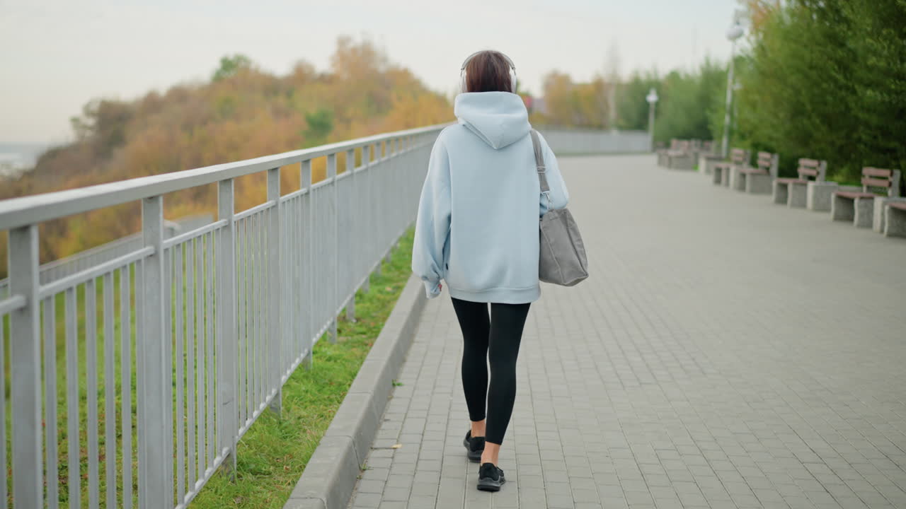 Tampak belakang seorang wanita atletis mengenakan hoodie biru dan legging hitam berjalan di sepanjang pagar besi dalam suasana cerah dan tenteram. Kebugaran luar ruangan, gaya hidup sehat, jalan pagi di alam.