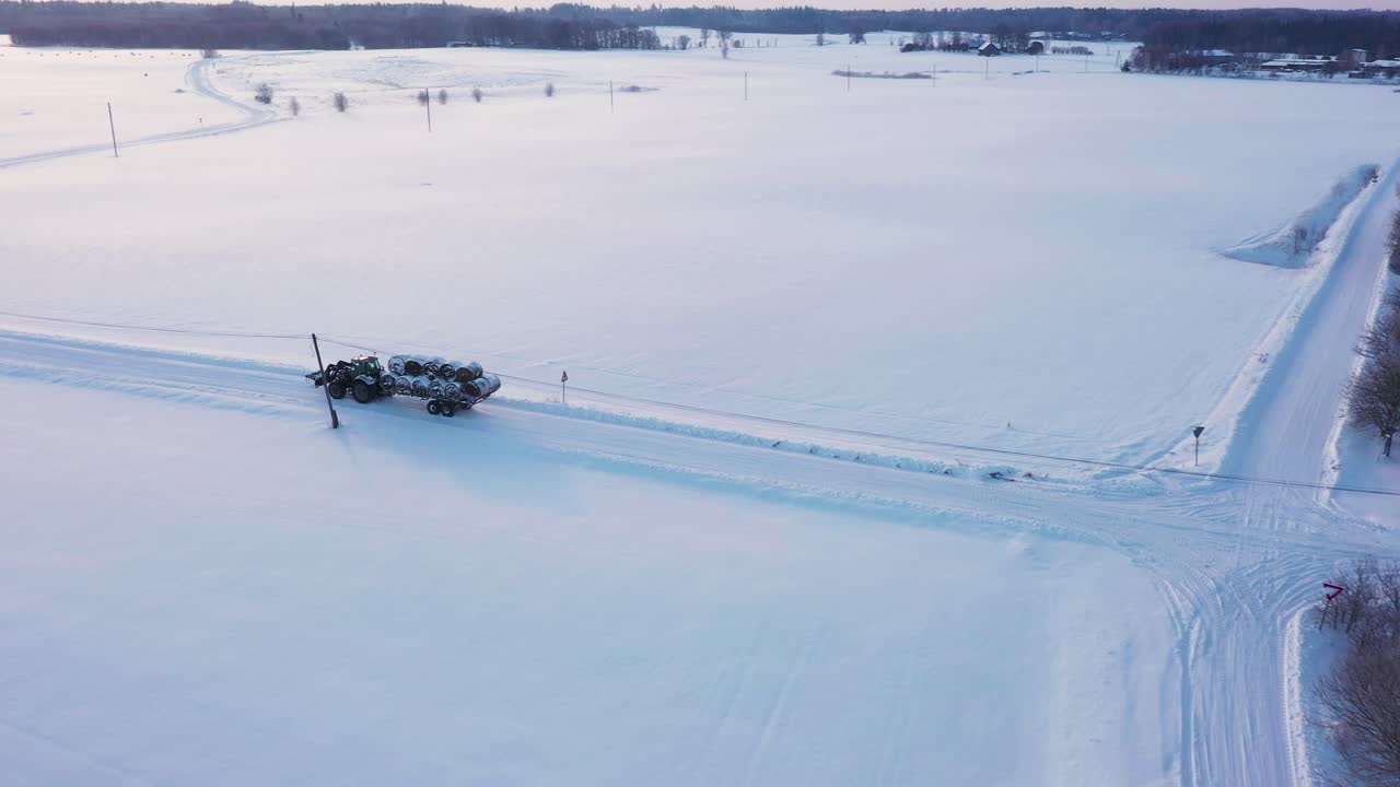 vista aérea del remolque del tractor que lleva fardos enrollados a través del campo agrícola rural de invierno nevado