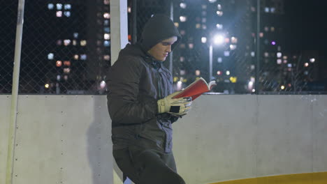 man in hoodie leaning against metallic goalpost reading book outdoors at night under urban city lights, creating a thoughtful and reflective scene of solitude