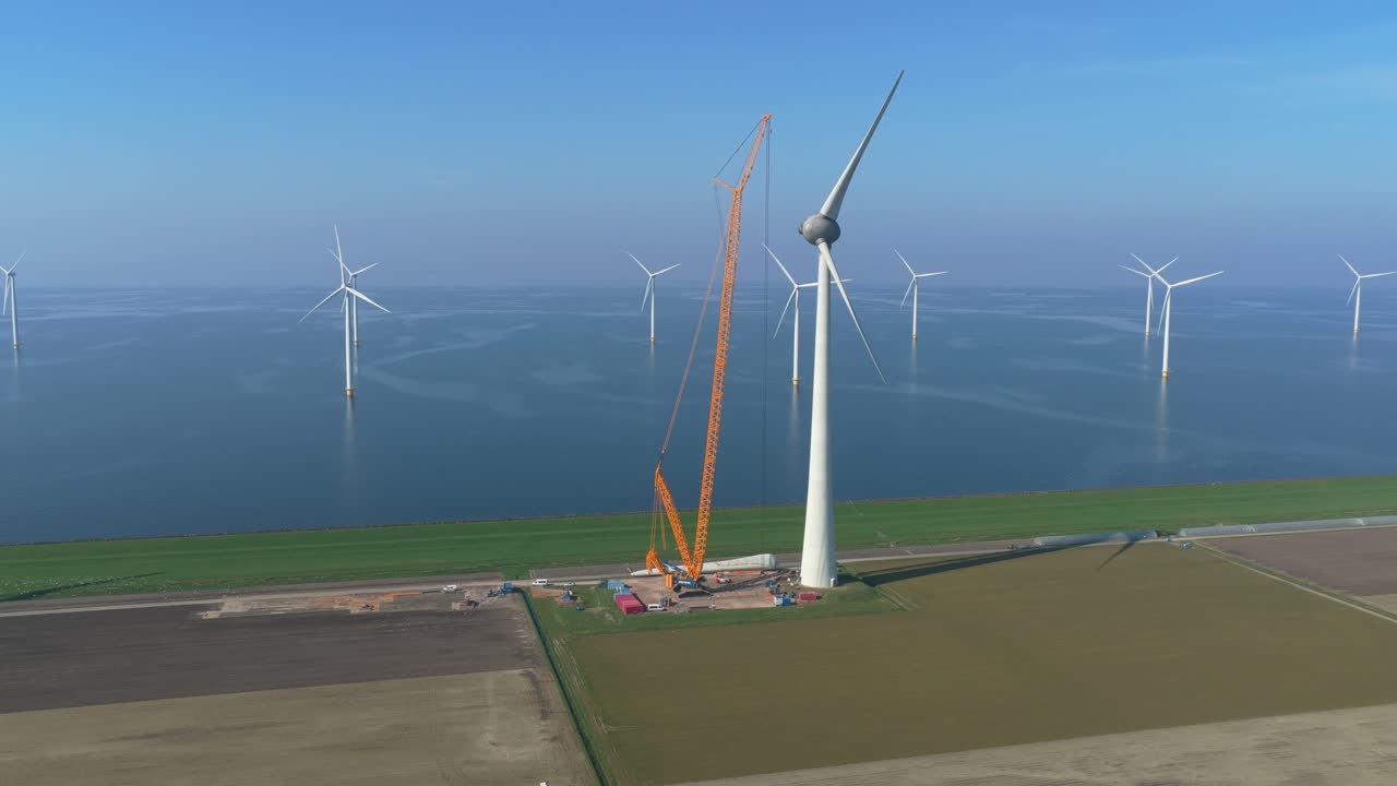 Vista aerea che mostra una grande gru che solleva una pala enorme per assemblare una turbina eolica sulla costa, con un parco eolico offshore sullo sfondo.