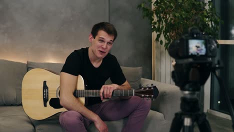 handsome caucasian man in black t shirt making video blog about musical instruments or recording the song for his followers while sitting on a couch at home. recording camera on a foreground