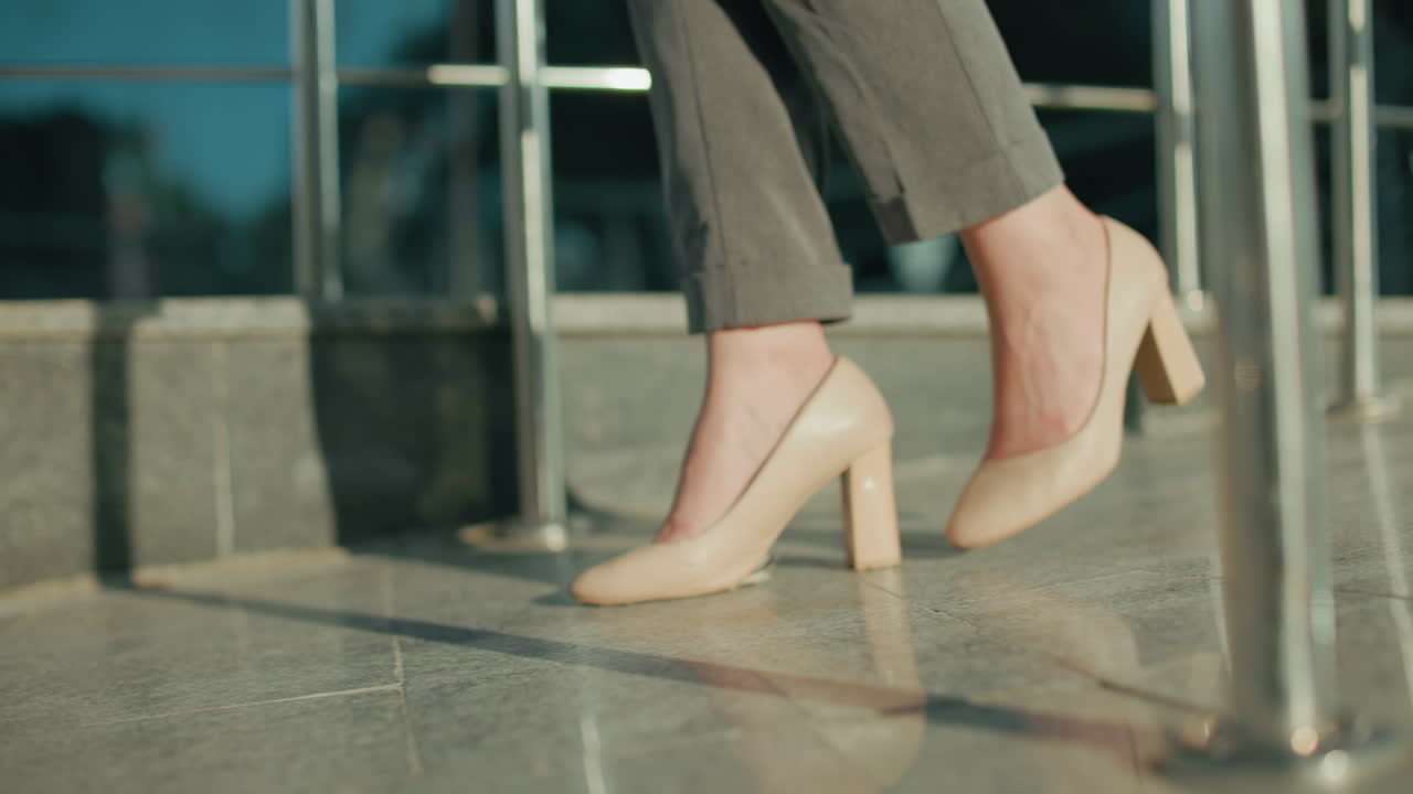 Leg view of woman in heels walking on shiny tiled floor, sunlight casting clear reflections and shadows, dressed in formal trousers in professional setting with metal railings and polished walkway