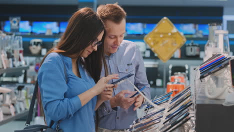 a young man and woman hold in their hands two smartphones standing around a showcase with smartphones in an electronics store choosing from two the best. comparison of two devices. large selection of products