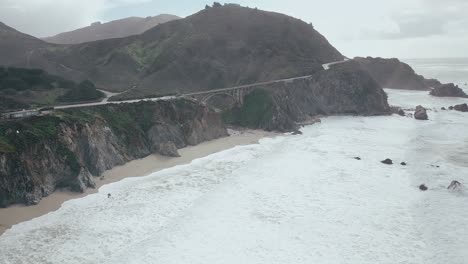 Amplia-Vista-Aérea-De-Drones-Que-Se-Dirige-A-Lo-Largo-De-La-Costa-De-La-Autopista-Del-Pacífico-Sur-Para-Terminar-En-Una-Vista-Cercana-Del-Tráfico-En-Arco-Que-Cruza-El-Puente-Del-Terreno-Montañoso