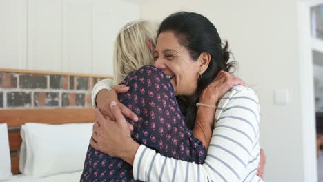 Two-happy-diverse-senior-women-embracing-and-laughing-in-sunny-bedroom,-slow-motion