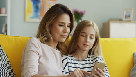 cute little girl with her mother scrolling on the smartphone and talking while sitting on sofa in living room