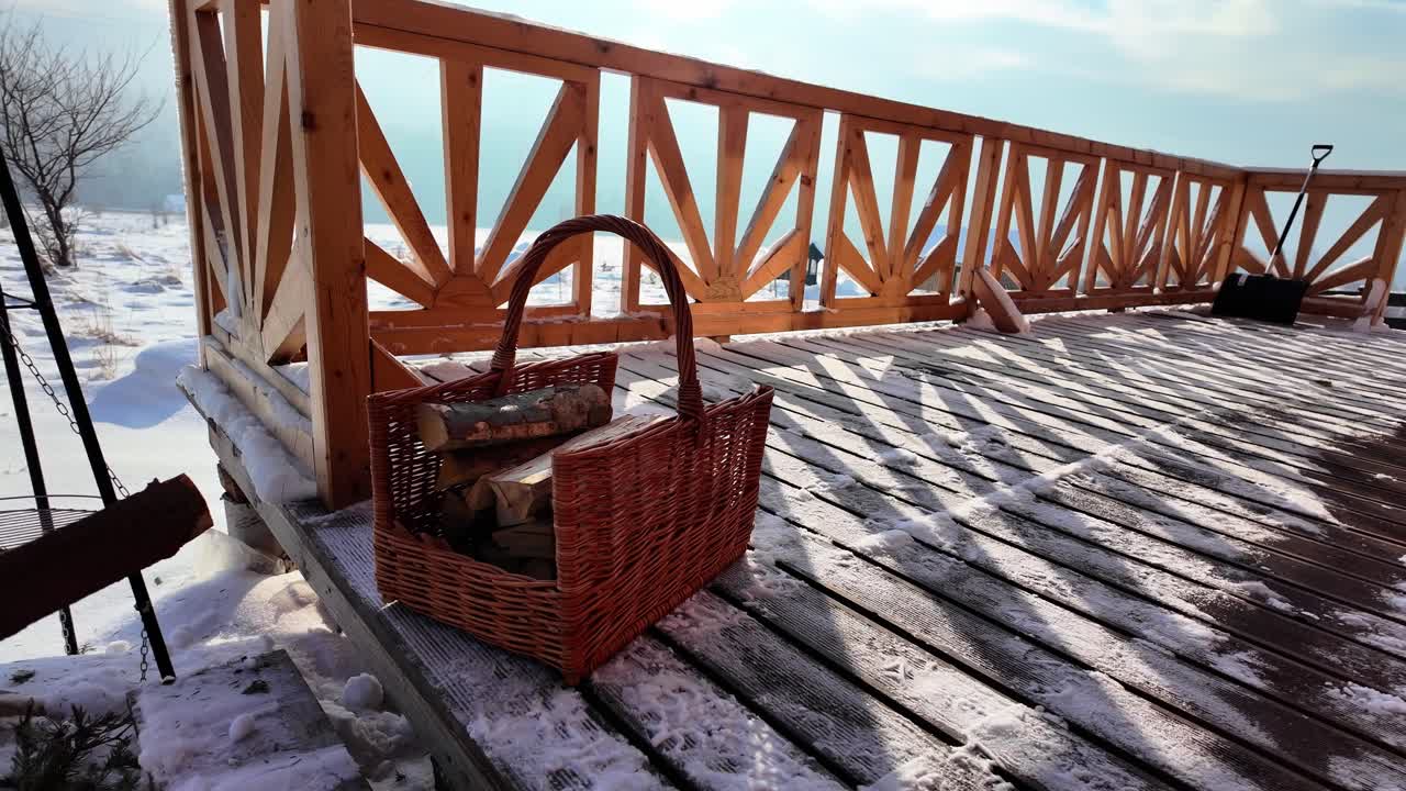 Una mano enguantada arroja trozos de madera a una cesta en una terraza iluminada por el sol. La nieve, una pala y un cielo nublado crean un ambiente invernal natural.