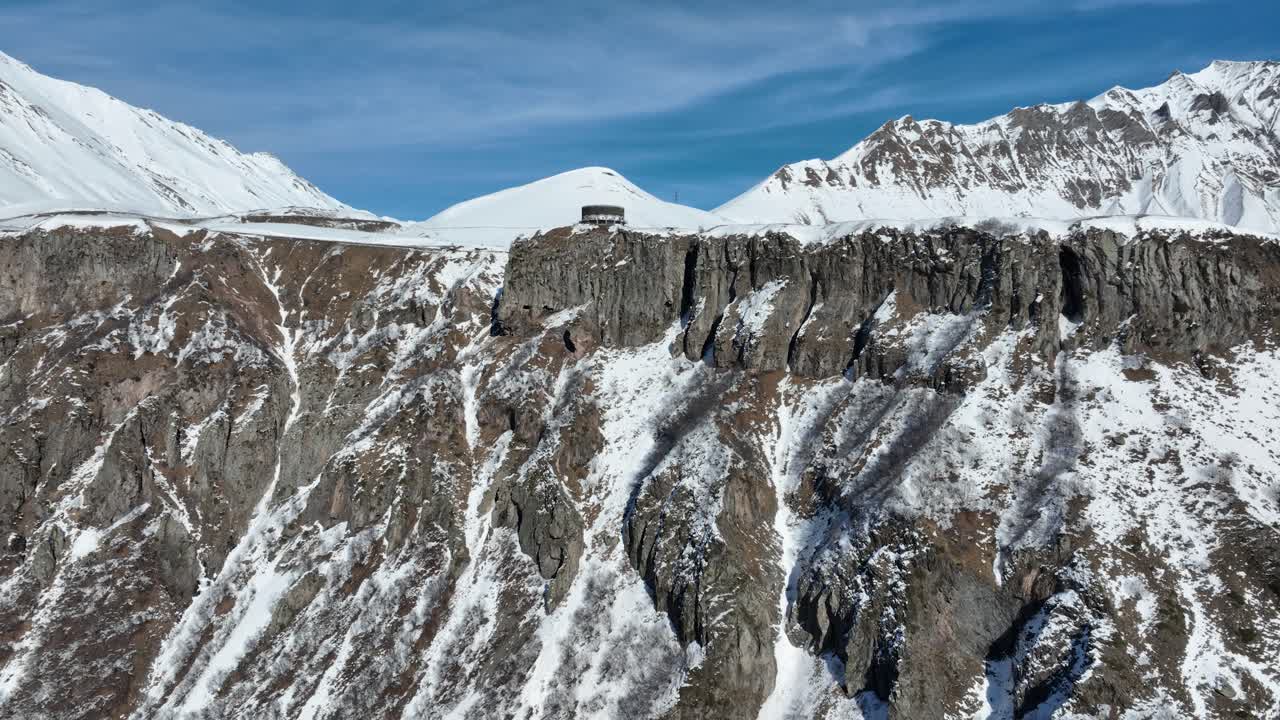 A dramatic cliffside road with a stunning drop below, offering a breathtaking journey through high-altitude snowy peaks. The rugged rock face contrasts beautifully with the soft white snow
