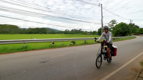 Female-bike-rider-passing-by-while-riding-bicycle-on-countryside-paved-road,-Thailand