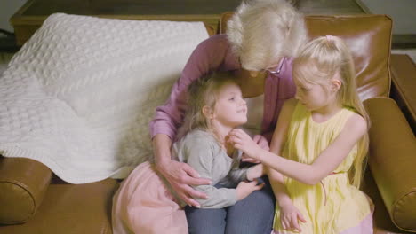 Top-View-Of-Grandmother-Hugging-To-Her-Two-Granddaughters-Sitting-On-The-Sofa-While-Talking-In-The-Living-Room-At-Home