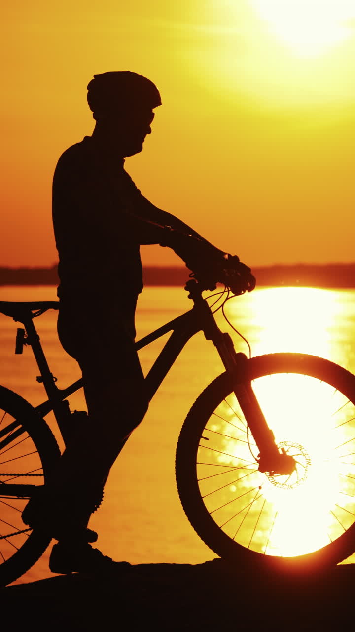 Silhouette of a cyclist near the river at sunset. Sporting man in helmet and glasses rides his bike on the background of beautiful sunset over the water surface. Vertical video
