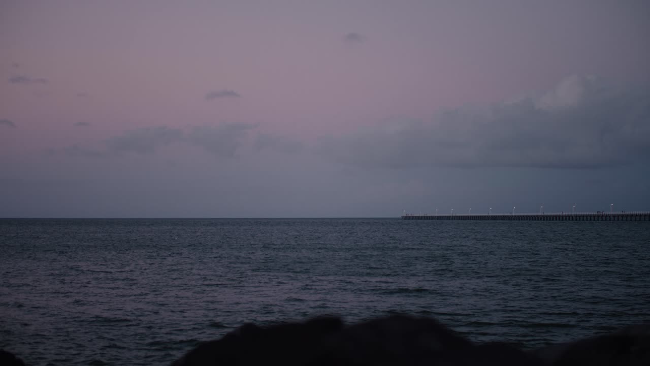 해가 천천히 지면서 광활한 바다로 뻗어나가는 긴 부두의 저속 촬영, 멋진 파스텔 핑크색과 푸른 하늘이 어둠으로 바래다
