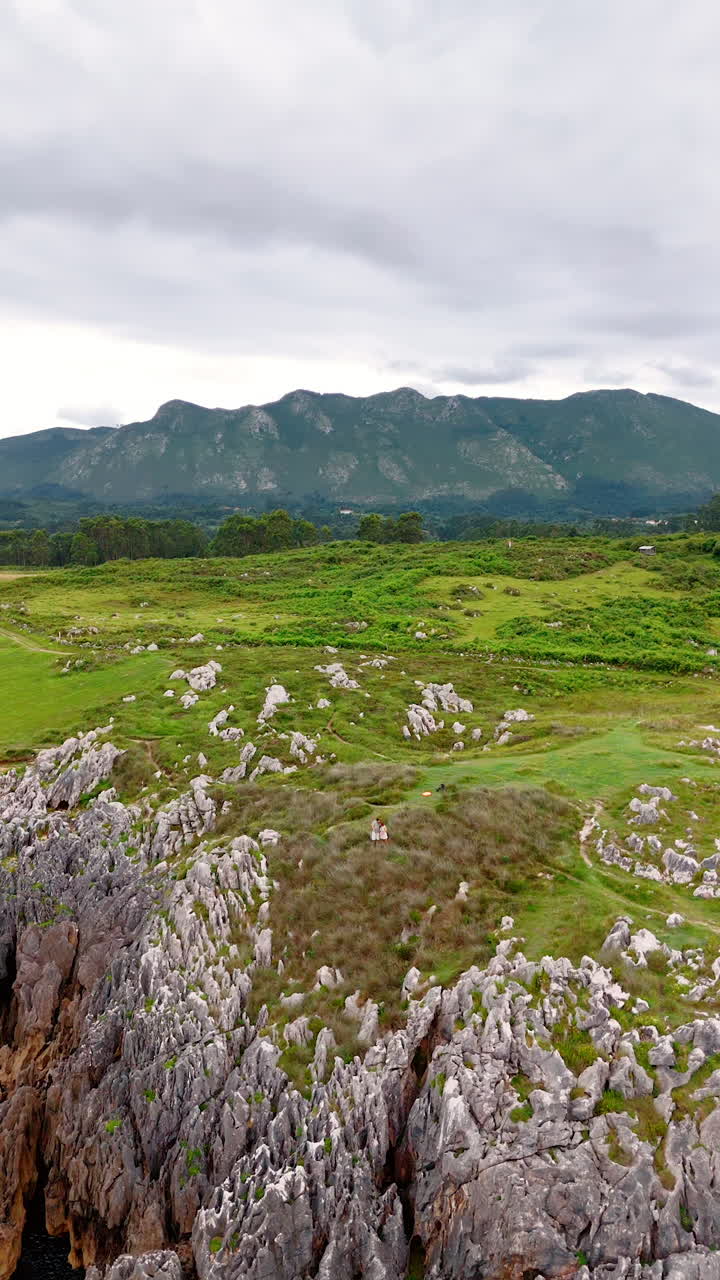 Couple stands at the rocky cliff on the sea shore. Drone footage around the mountainous coast with spiky stones. Mountain range under grey sky at backdrop. Vertical video.