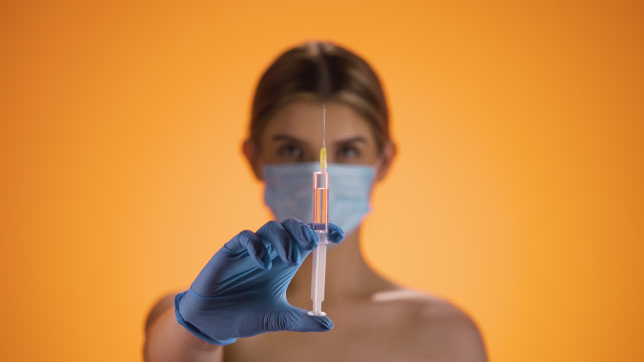 toma media de una joven enfermera bonita que lleva una máscara ffp2 mientras sostiene una jeringa con líquido naranja en la cámara para vacunar contra enfermedades o virus de la corona frente a un fondo naranja