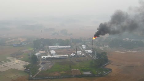 Aerial-view-establishing-Petrochemical-refinery-plant-flaming-smoking-flare-stack-over-misty-agricultural-farmland-at-sunrise