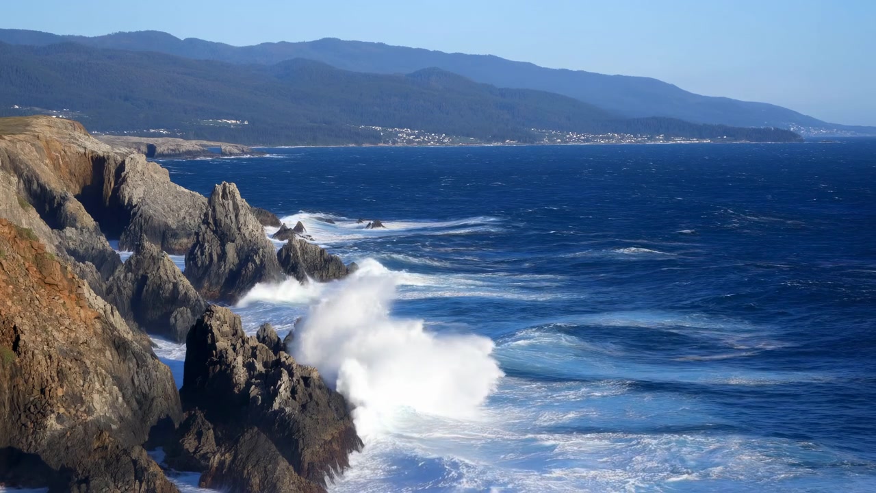 La spuma schiumosa si riversa nelle scure rocce costiere ed esplode verso l'alto in mutevoli pennacchi, con scogliere illuminate dal sole a sinistra, mare aperto a destra e montagne stratificate che delineano l'orizzonte lontano.
