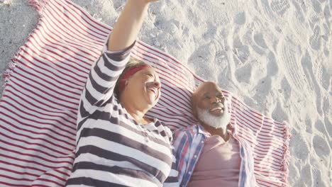 una feliz pareja afroamericana de edad avanzada acostada en una manta en la playa, cámara lenta