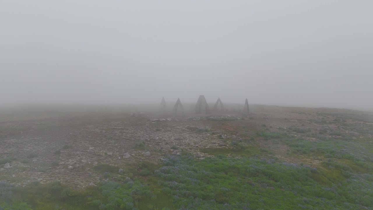 Vista aerea con drone che mostra un misterioso cerchio di sculture che emerge attraverso una fitta nebbia in un paesaggio brullo, creando una scena suggestiva e d'atmosfera nel remoto territorio islandese.