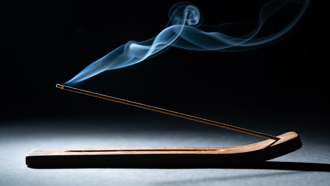 Varilla de incienso humeante emitiendo humo azul de una brasa caliente sobre una mesa con un soporte de madera, formando rizos.