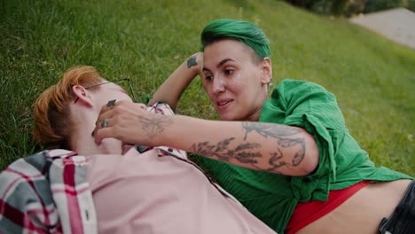 Close-up-shot-of-a-girl-with-a-short-haircut-with-green-hair-in-a-checkered-green-shirt-stroking-her-girlfriend-a-blonde-with-a-short-haircut-wearing-glasses-in-a-checkered-pink-shirt-during-her-date-in-the-park-lying-on-the-lawn-on-green-grass-in-the-summer