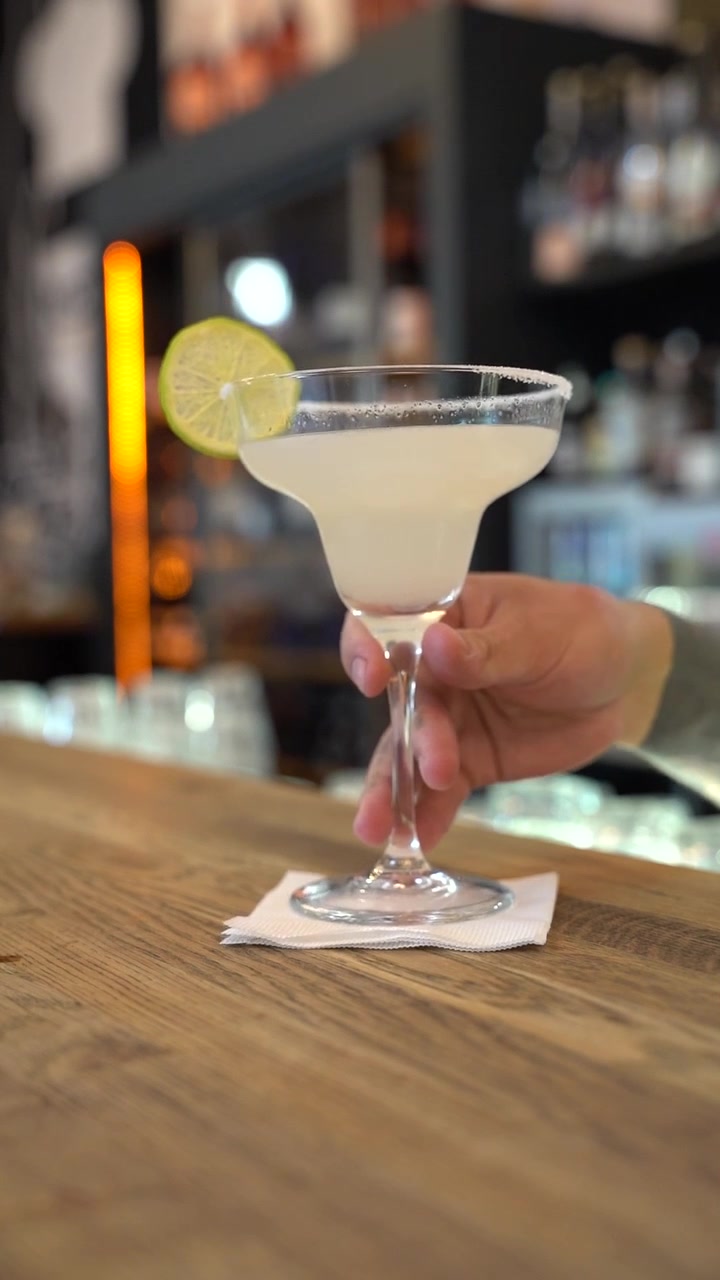 Uma margarita refrescante com borda de sal e guarnição de limão repousa elegantemente em um balcão de bar de madeira. A iluminação suave do bar e as garrafas ao fundo criam uma atmosfera aconchegante e convidativa.