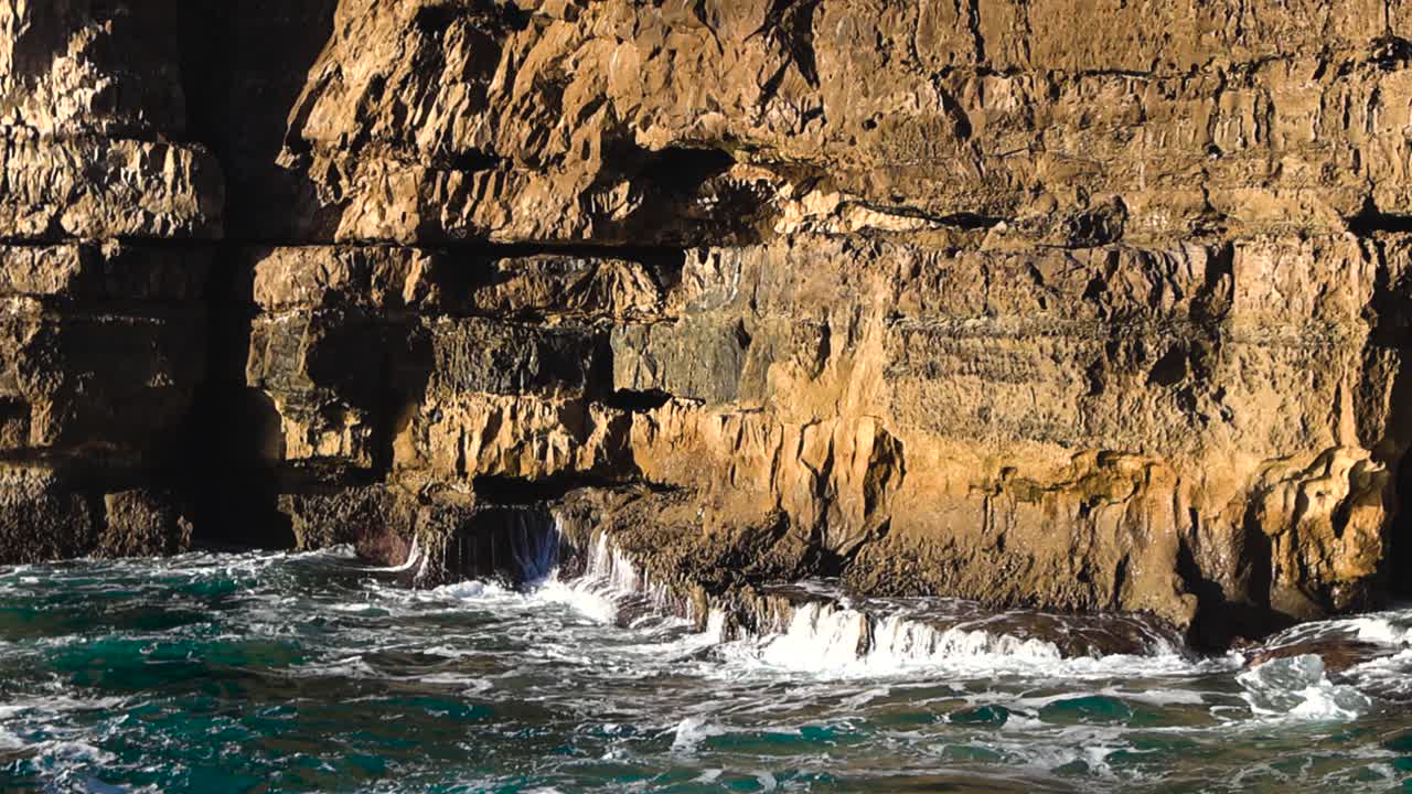 Powerful Atlantic waves crash into the dramatic Boca do Inferno sea cave, sending white foam and spray against sunlit yellow cliffs in a wild coastal scene