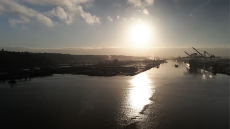 Bright-golden-sun-reflecting-off-calm-water,-approaching-an-industrial-port-operation,-aerial-establish